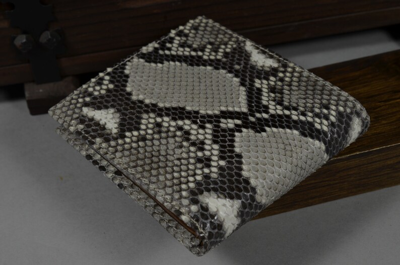 May include: A bi-fold wallet crafted from genuine snakeskin. The wallet displays a pattern of grey, black, and white scales. The rectangular wallet appears to be unused.