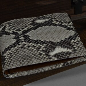 May include: A snakeskin bi-fold wallet. The wallet has a pattern of grey, black, and white scales. The interior is brown leather. The wallet is on a wooden surface.