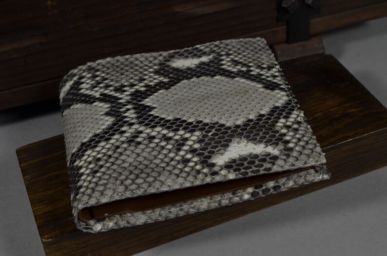 May include: A snakeskin bi-fold wallet. The wallet has a pattern of grey, black, and white scales. The interior is brown leather. The wallet is on a wooden surface.