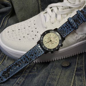 Könnte beinhalten: Ein blaues und schwarzes Uhrenarmband mit weißem Zifferblatt und schwarzen Zeigern. Die Uhr befindet sich auf einem weißen Sneaker mit blauem Jeans-Hintergrund.