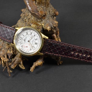 Puede incluir: Una correa de reloj de cuero marrón con un patrón de cocodrilo. La correa está unida a un reloj dorado con una esfera blanca y agujas negras. La esfera del reloj tiene una ventana de fecha y un indicador de 24 horas.