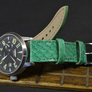 以下が含まれることがあります： 銀色のバックルが付いた緑色の革製の腕時計のストラップ。ストラップは、蛇のようなテクスチャパターンになっています。時計の文字盤は黒で、白い数字と針が付いています。時計の文字盤には、「АВИАТОР」と「17 Jewels」の文字も書かれています。