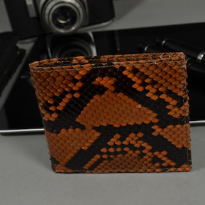 Op de afbeelding: Een bruine en zwarte slangenleren portemonnee. De portemonnee is gesloten en heeft een textuur.