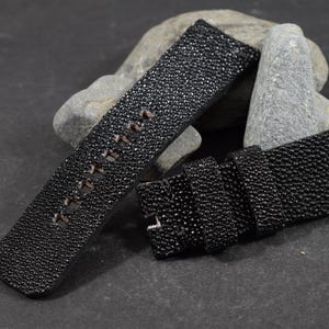 Op de afbeelding: Tweedelig zwart horlogebandje met een getextureerd, gespikkeld oppervlak. De band heeft bruine stiksels en een zilveren gesp. De horlogeband wordt getoond op een donkergrijze ondergrond met grijze stenen op de achtergrond.