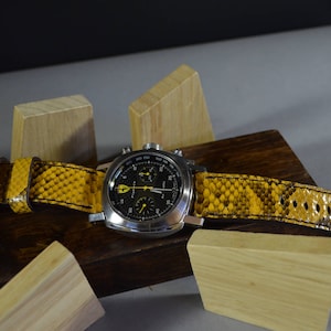 Könnte beinhalten: Eine schwarze Chronographenuhr mit einem gelben Lederarmband. Das Armband hat ein strukturiertes Muster, das an Schlangenhaut erinnert. Die Uhr liegt auf einer Holzoberfläche.