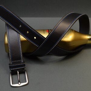 Può includere: Una cintura in pelle nera con cuciture blu e beige avvolta attorno a una bottiglia dorata. La cintura ha una fibbia argentata.