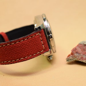 May include: A stainless steel watch with a red canvas strap and cream stitching. A small, reddish-brown rock sits to the right of the watch. The watch is on a light tan surface.