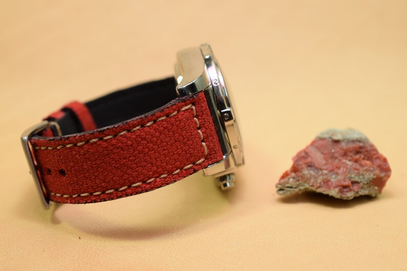 May include: A stainless steel watch with a red canvas strap and cream stitching. A small, reddish-brown rock sits to the right of the watch. The watch is on a light tan surface.