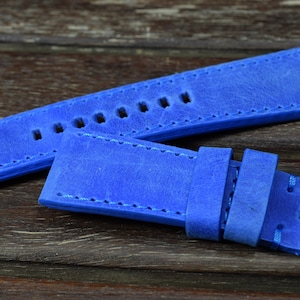 May include: A blue leather watch strap with stitching around the edges. The strap has multiple holes for adjusting the size.