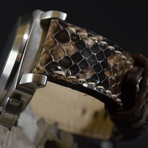May include: A stainless steel wristwatch with a snakeskin leather band. The band features a pattern of brown, tan, and black scales. The watch has a round face and a braided leather loop.