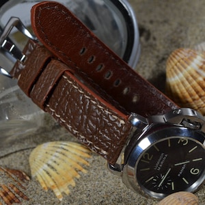May include: A brown leather watch strap with a textured surface and white stitching. The strap is attached to a silver watch with a black face and white markings. The watch face reads "Luminor Marina Panerai".