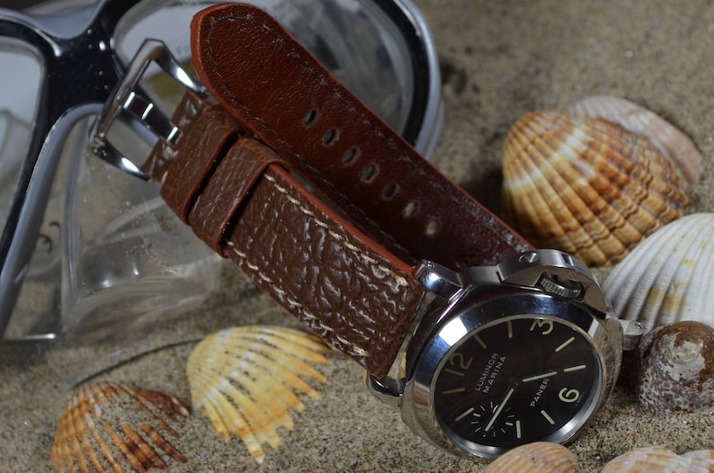 May include: A brown leather watch strap with a textured surface and white stitching. The strap is attached to a silver watch with a black face and white markings. The watch face reads "Luminor Marina Panerai".