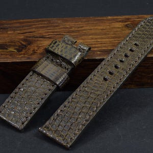 May include: A brown leather watch strap with a textured alligator pattern. The strap has stitching around the edges and a buckle on one end.
