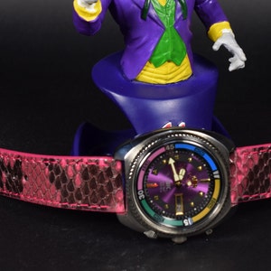 Puede incluir: Un reloj de pulsera morado y plateado con una correa de piel de serpiente rosa. La esfera del reloj tiene un bisel colorido y una ventana de fecha.