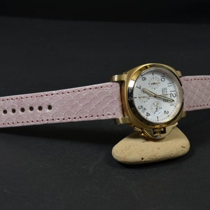 May include: A gold-tone wristwatch with a white face and black hands. The watch is on a pink leather strap with a crocodile pattern.
