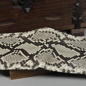 Pode incluir: Uma carteira de pele de cobra marrom claro com um padr&atilde;o preto e branco. A carteira est&aacute; dobrada ao meio e est&aacute; sobre uma superf&iacute;cie de madeira.