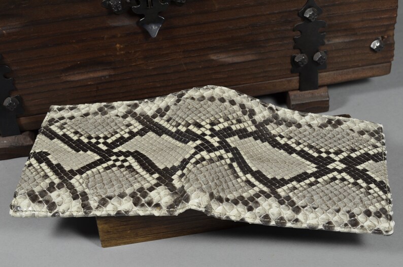 Pode incluir: Uma carteira de pele de cobra marrom claro com um padr&atilde;o preto e branco. A carteira est&aacute; dobrada ao meio e est&aacute; sobre uma superf&iacute;cie de madeira.