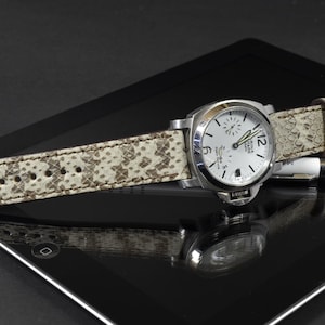 May include: A white watch with a silver band and a white face with black markings. The watch is on a white and brown patterned leather strap.