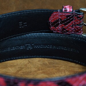 May include: A black leather belt with a red snakeskin pattern. The belt is handmade in Ubrique, Spain. The belt buckle is silver and has a simple design. The belt is 3.5 inches wide.