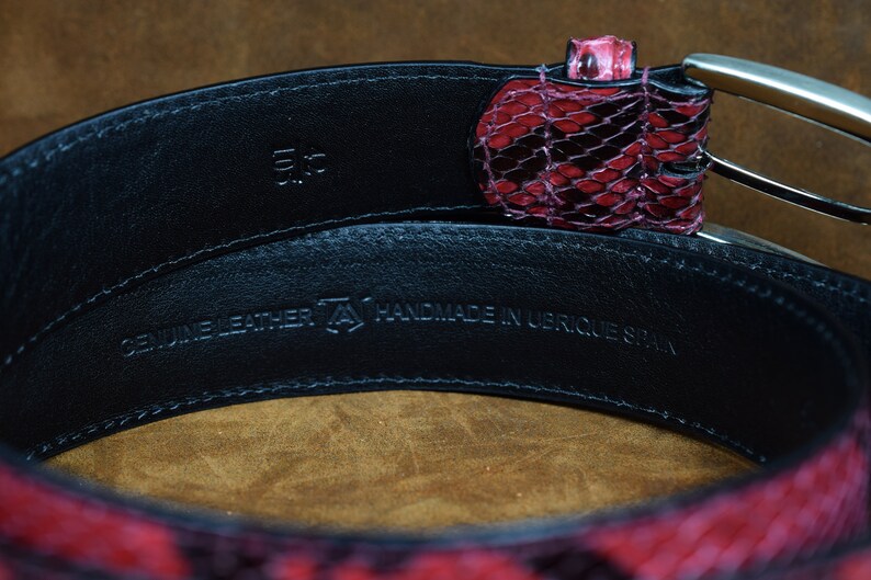 May include: A black leather belt with a red snakeskin pattern. The belt is handmade in Ubrique, Spain. The belt buckle is silver and has a simple design. The belt is 3.5 inches wide.