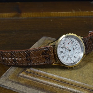 Peut inclure: Une montre-bracelet dorée avec un cadran blanc et des chiffres romains noirs. La montre a un bracelet en cuir marron avec un motif peau de serpent.