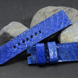 Op de afbeelding: Een helderblauwe horlogeband met een textuur, reptielachtig patroon. De band heeft meerdere gaten voor aanpassing en een gespsluiting. De horlogeband wordt getoond tegen een donkere achtergrond met grijze stenen.