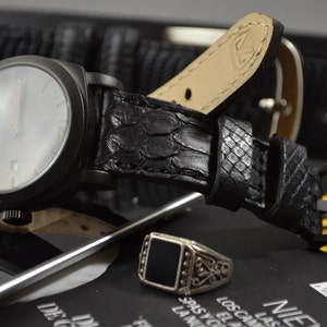 Op de afbeelding: Een zwart polshorloge met een witte wijzerplaat en een zwarte leren band. Het horloge ligt op een zwart oppervlak met een zilveren ring met een zwarte steen.