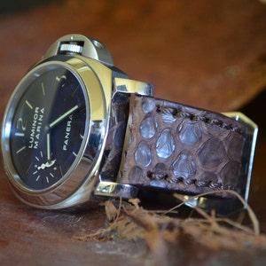 Może przedstawiać: Srebrny zegarek na rękę z czarną tarczą i brązowym skórzanym paskiem. Tarcza zegarka ma białe oznaczenia i tekst "Luminor Marina" oraz "Panerai".