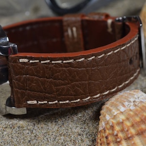 May include: A brown leather watch strap with white stitching. The strap has a textured surface and is shown on a background of seashells.