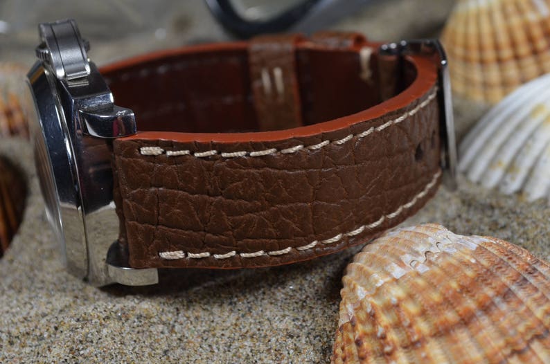 May include: A brown leather watch strap with white stitching. The strap has a textured surface and is shown on a background of seashells.