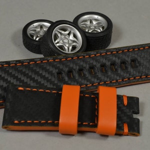 Peut inclure: Un bracelet de montre noir avec des coutures et des accents orange, présentant un motif de tissage en fibre de carbone. Trois roues de voiture miniatures avec des jantes argentées sont positionnées au-dessus du bracelet.