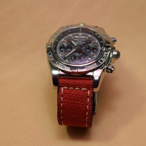 May include: A stainless steel Breitling watch with a dark gray face and a red canvas strap. The watch features multiple subdials and a bezel with engraved markings. The red strap is stitched with white thread.
