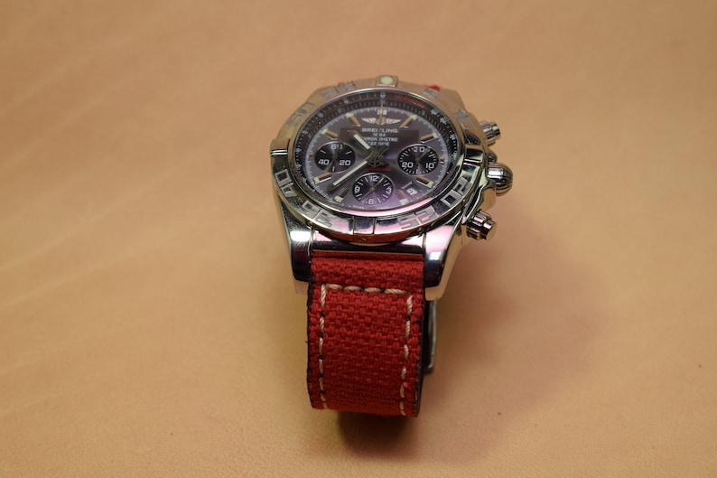 May include: A stainless steel Breitling watch with a dark gray face and a red canvas strap. The watch features multiple subdials and a bezel with engraved markings. The red strap is stitched with white thread.
