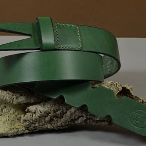 Può includere: Una cintura in pelle verde con un design di fibbia unico. La cintura è piegata e appoggiata su un pezzo di legno.