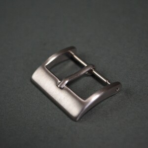 May include: A silver-colored watch buckle with a rectangular shape and a curved design. The buckle has a bar for the watch strap and a pin to secure the strap. The background is a dark gray color.