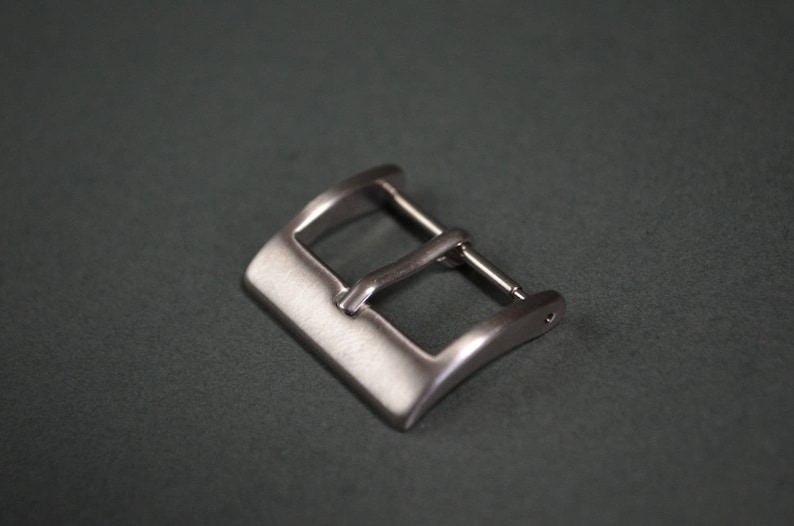 May include: A silver-colored watch buckle with a rectangular shape and a curved design. The buckle has a bar for the watch strap and a pin to secure the strap. The background is a dark gray color.