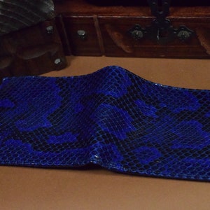 Peut inclure: Un portefeuille en cuir de serpent bleu avec un motif noir. Le portefeuille est pli&eacute; en deux et repose sur une surface brune.