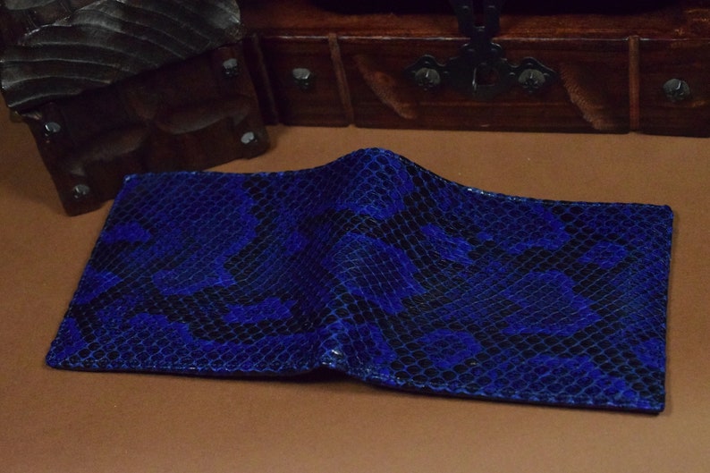 Peut inclure: Un portefeuille en cuir de serpent bleu avec un motif noir. Le portefeuille est pli&eacute; en deux et repose sur une surface brune.