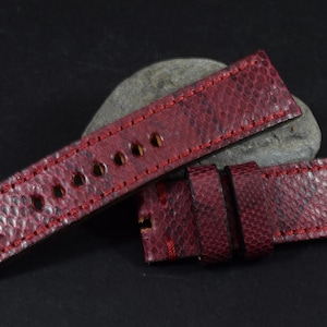 Può includere: Un cinturino da orologio in pelle rossa con una superficie testurizzata e cuciture rosse. Il cinturino è piegato a metà e si trova su una roccia grigia.