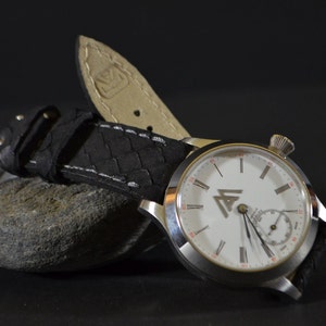 Puede incluir: Un reloj de pulsera plateado con una esfera blanca y una correa de cuero negra. La esfera del reloj tiene marcadores de hora en números romanos y un pequeño dial de segundos. El reloj tiene un logotipo con las letras "AI" en un triángulo.