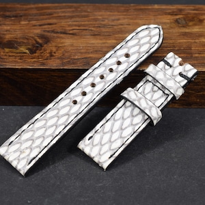 Può includere: Due cinturini per orologio con motivo a scaglie bianche e grigie e cuciture nere. I cinturini sono realizzati in un materiale strutturato e sono esposti su una superficie di legno scuro. Un accessorio elegante.