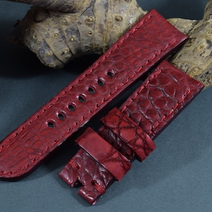 May include: Red leather watch strap with a crocodile pattern. The strap has stitching around the edges and multiple holes for adjusting the size.