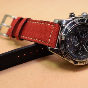 May include: A stainless steel chronograph watch with a dark face and a red suede strap. The watch features multiple dials and a silver buckle. The strap is stitched with white thread.