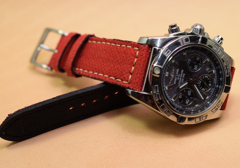 May include: A stainless steel chronograph watch with a dark face and a red suede strap. The watch features multiple dials and a silver buckle. The strap is stitched with white thread.