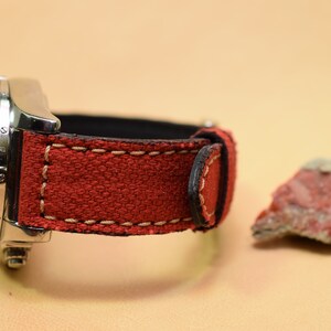 May include: A stainless steel watch with a red fabric band and cream stitching. The watch band is next to a small, reddish-brown rock. The watch has a black interior.