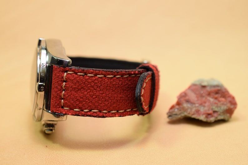 May include: A stainless steel watch with a red fabric band and cream stitching. The watch band is next to a small, reddish-brown rock. The watch has a black interior.