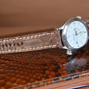 Può includere: Un orologio da polso con cassa color argento e quadrante bianco, con numeri e lancette nere. L'orologio ha un cinturino in pelle marrone con un motivo strutturato. È visibile anche una penna d'argento.