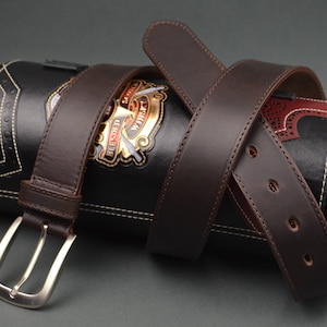 Könnte beinhalten: Ein dunkelbrauner Ledergürtel mit silberner Schnalle ist um eine zylindrische schwarze Ledertasche gewickelt. Die Tasche hat weiße Nähte und ein goldenes Emblem. Der Gürtel hat mehrere Löcher zur Anpassung.