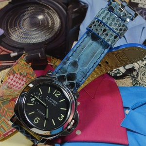 Könnte beinhalten: Eine Edelstahl Panerai Luminor Marina Uhr mit schwarzem Zifferblatt und leuchtenden Zeigern und Ziffern. Die Uhr hat ein blaues Lederarmband mit Schlangenmuster. Der Hintergrund umfasst bunte Stoffe und Kleidung.