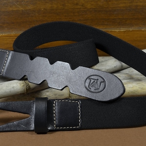 Könnte beinhalten: Ein schwarzer Ledergürtel mit einer Schnalle mit einem stilisierten Buchstaben "W". Der Gürtel ist aus gewebtem Stoff und hat einen genähten Rand.